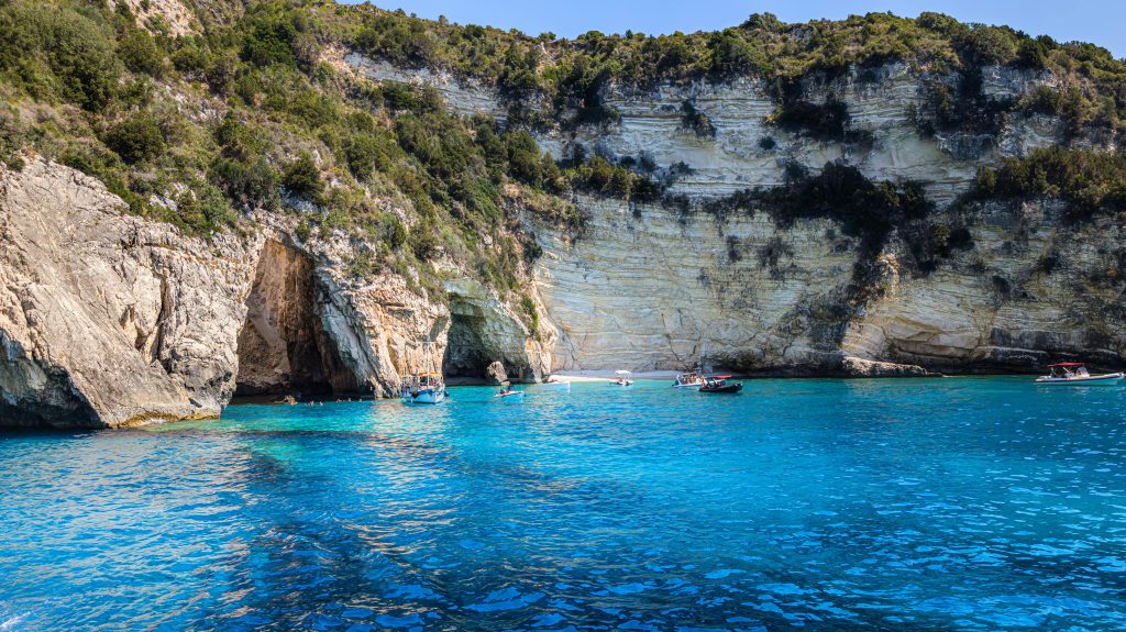 Small boats floating in bright turquoise water near Blue Cave and limestone cliffs in Paxos, Greece.